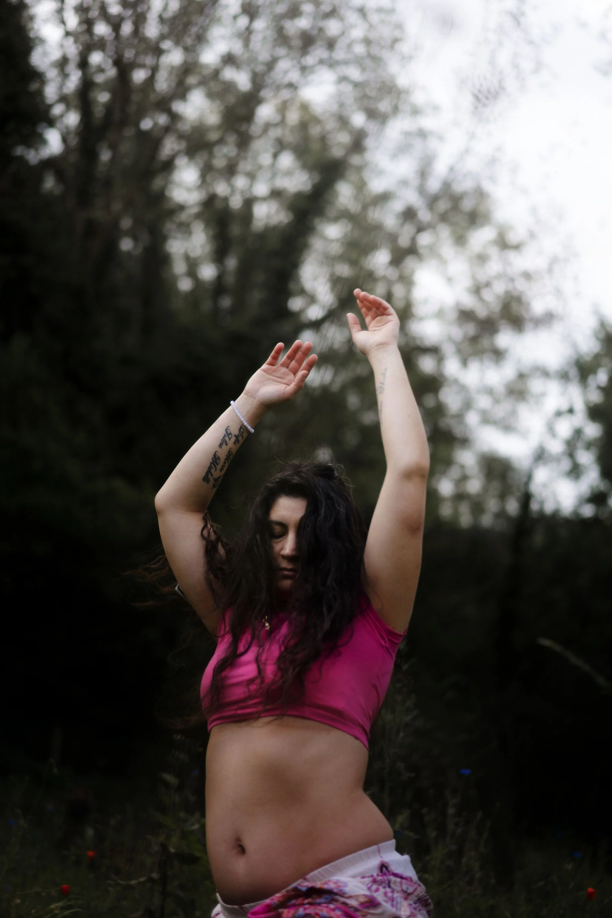 Une femme avec des tatouages sur les bras, portant un haut rose vif, danse ou pose avec les bras levés dans un environnement extérieur avec des arbres en arrière-plan.
