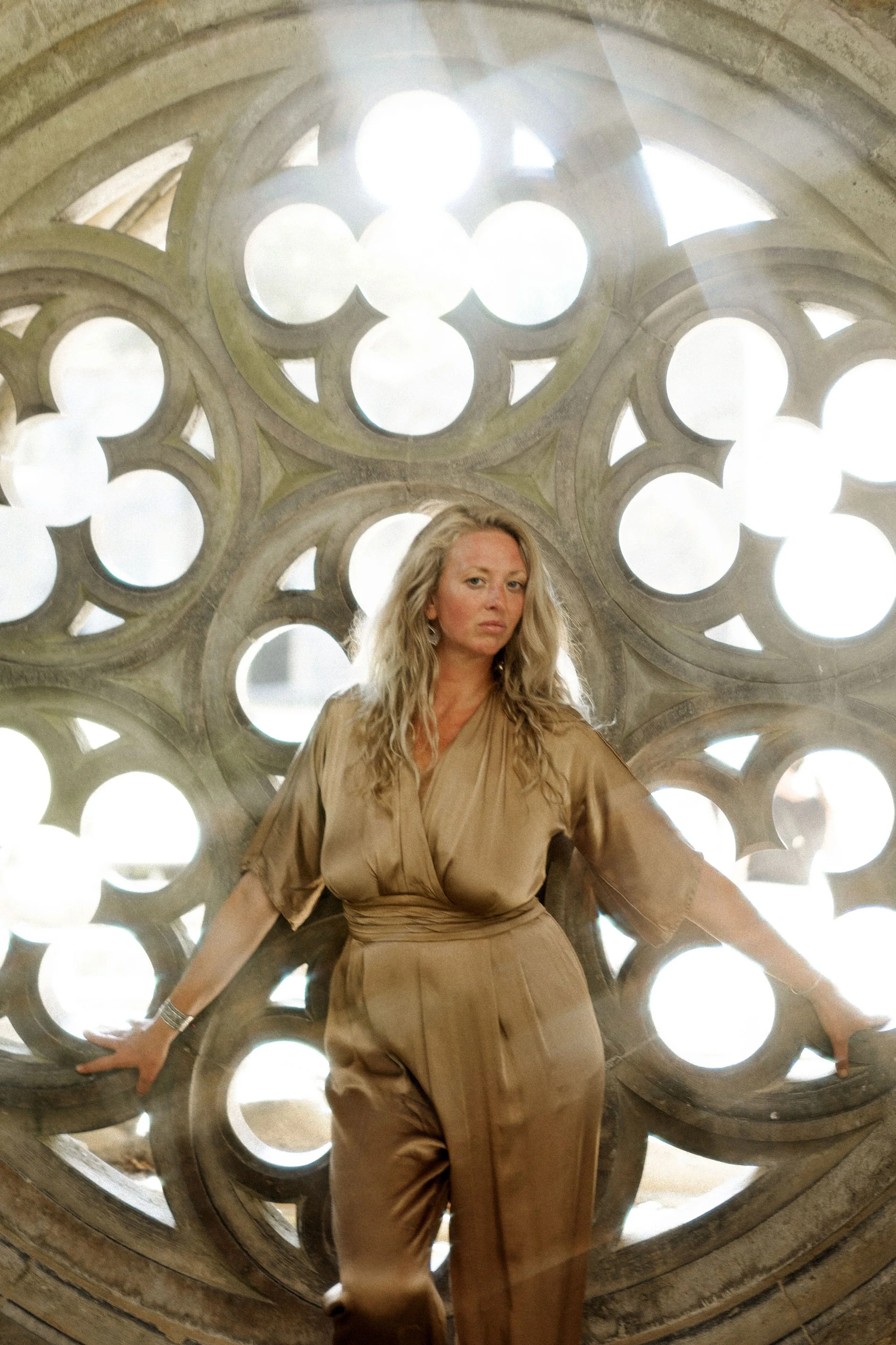 Une femme en tenue beige posant devant une structure architecturale avec des cercles et des arches, laissant entrer la lumière.