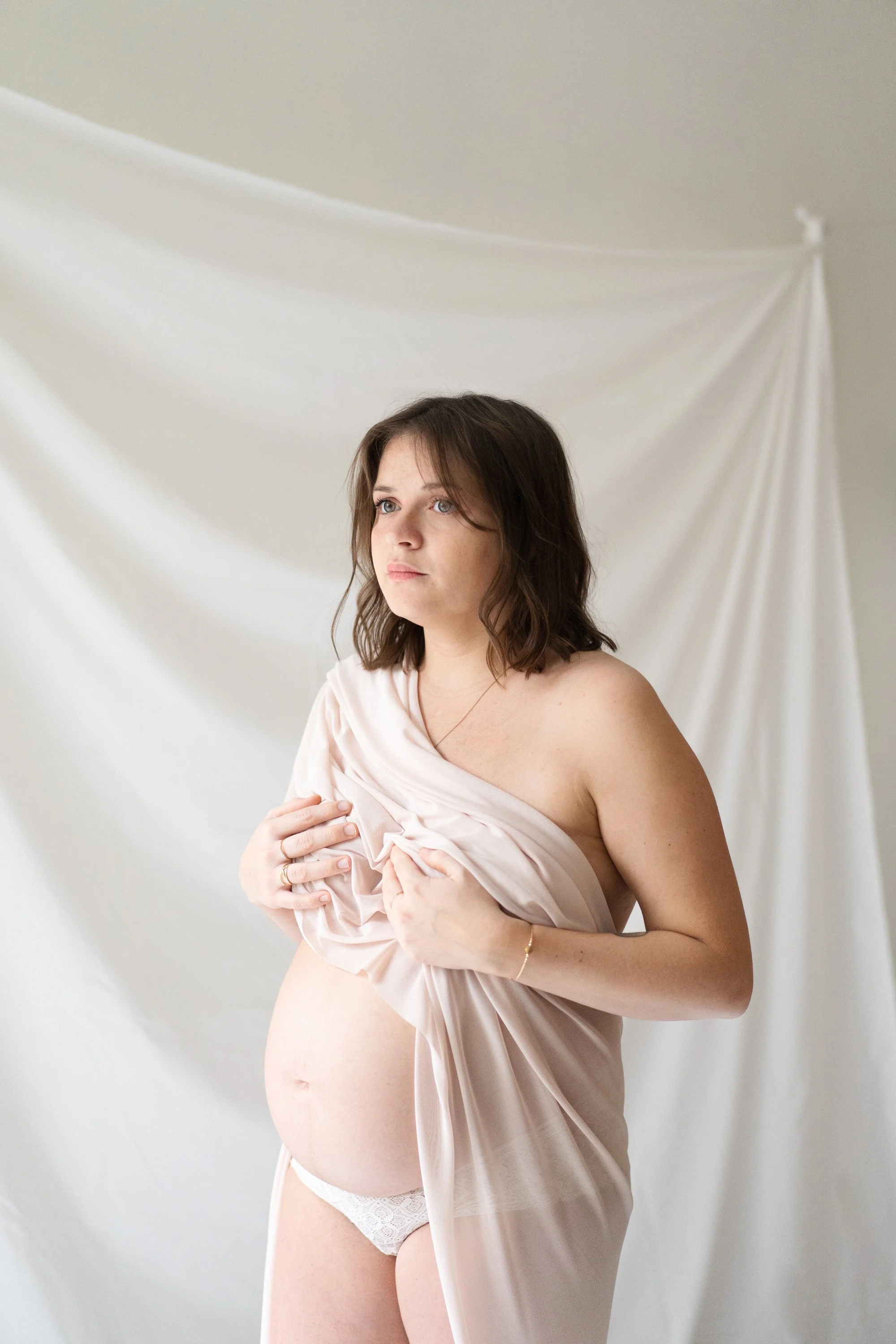 Une femme enceinte pose devant un fond blanc enroulé dans un tissu beige.