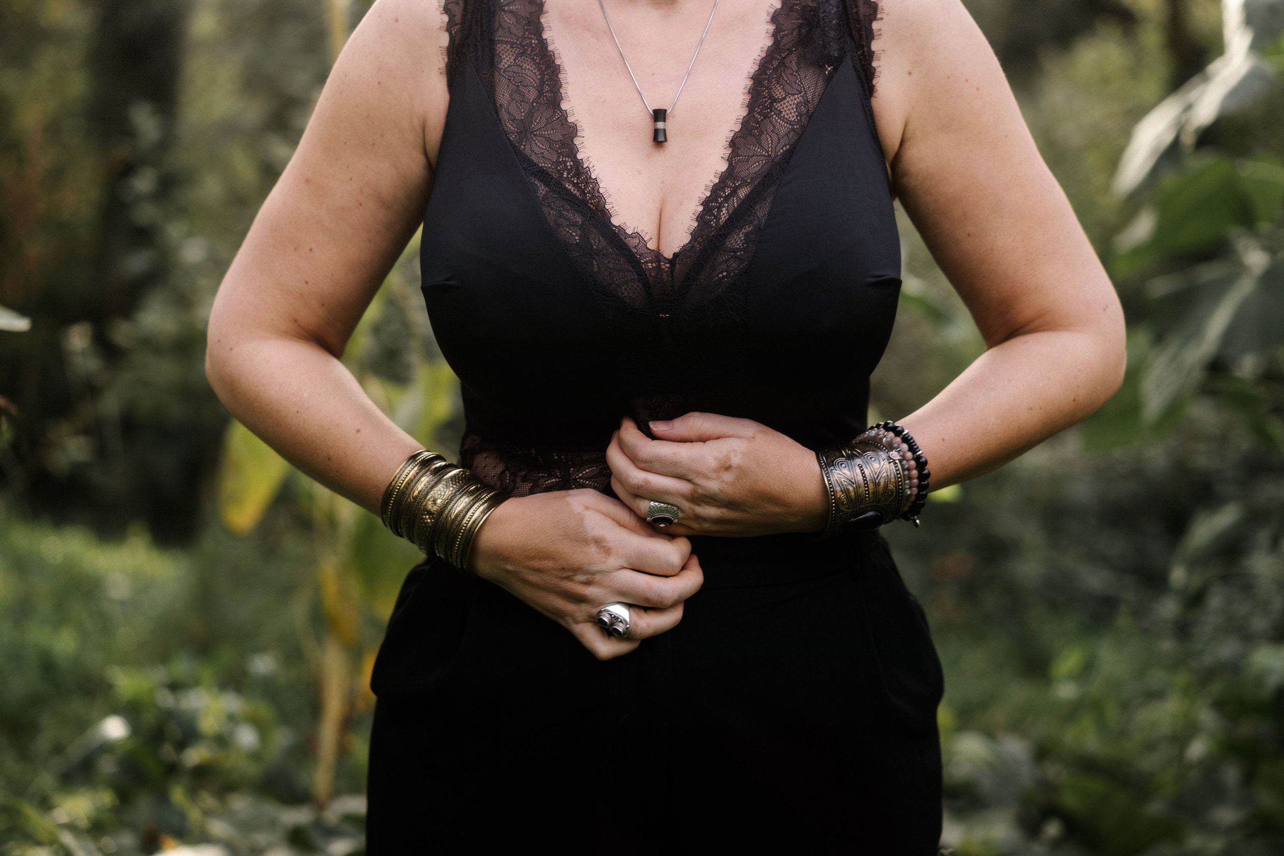 Femme portant un haut noir avec des détails en dentelle, portant plusieurs bracelets en métal, un collier avec un pendentif noir, les mains jointes au ventre, dans un environnement naturel avec des arbres en arrière-plan.