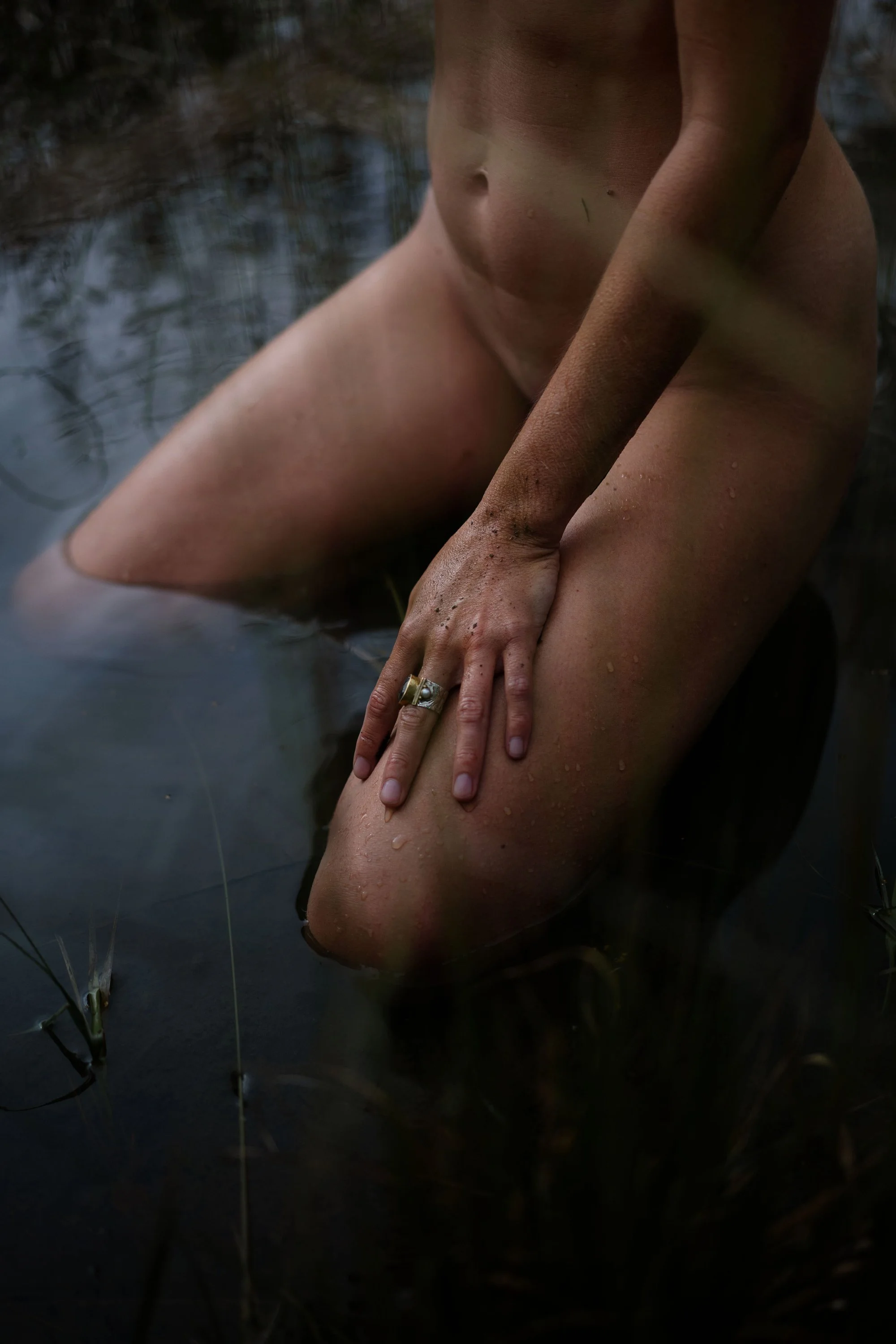 Une personne nue, assise dans l'eau, avec la main sur la cuisse, portant une bague, dans un environnement naturel humide. La photo est sombre et artistique.