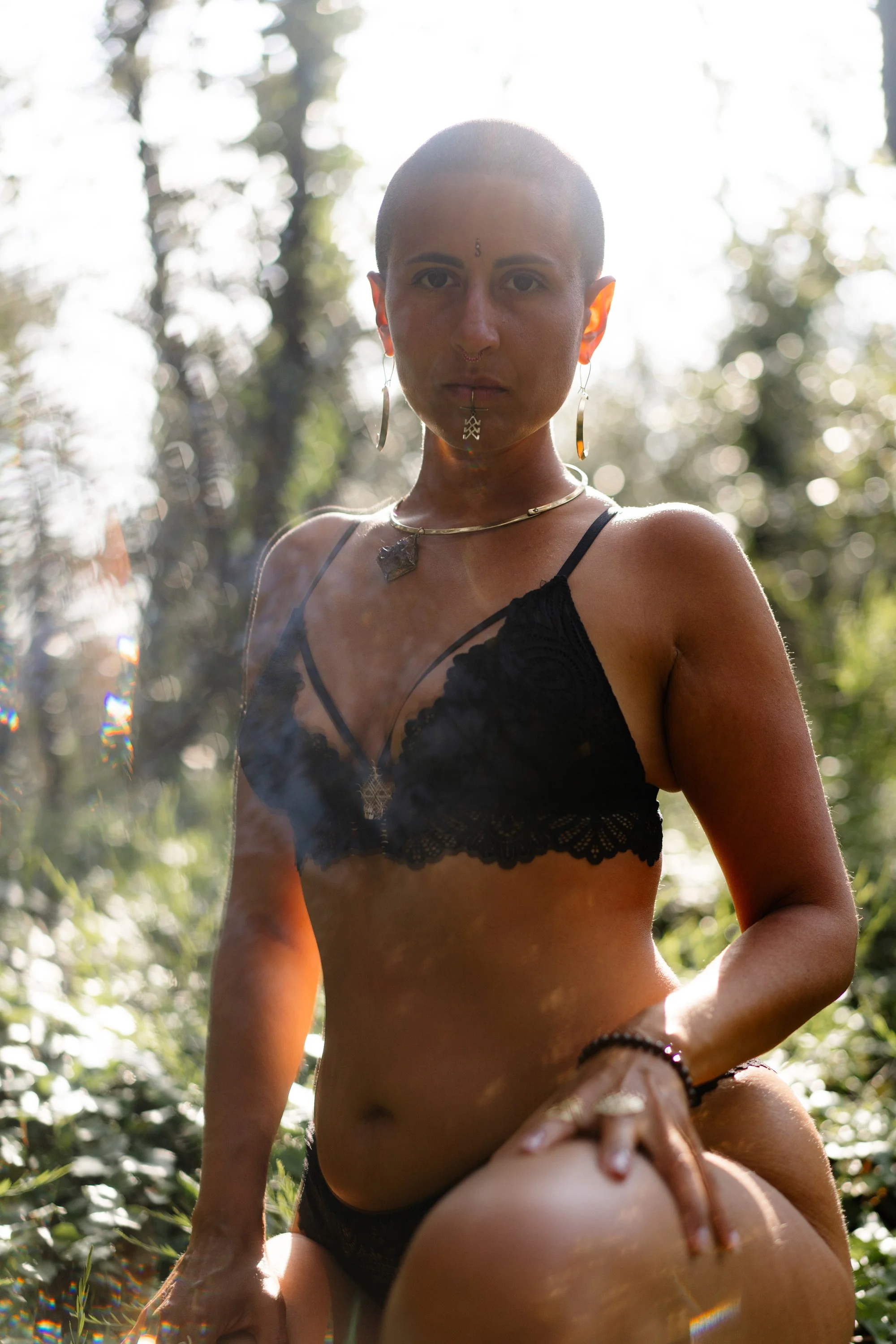 Femme nue avec des piercings, portant des bijoux, dans une forêt en plein jour, avec un arrière-plan bokeh de feuilles et de lumière solaire, posant de manière artistique.