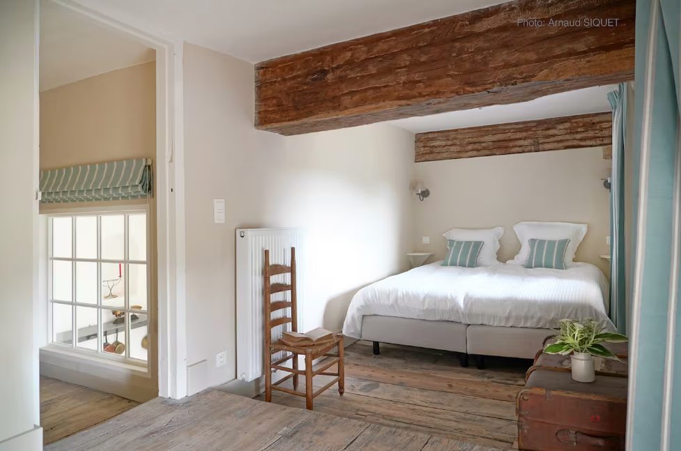 Chambre avec un lit blanc, deux oreillers à rayures vertes, murs blancs, poutres en bois au plafond, petite table de nuit, fauteuil en bois, plante en pot, fenêtre avec rideau bleu et fenêtre à carreaux dans une autre pièce visible.