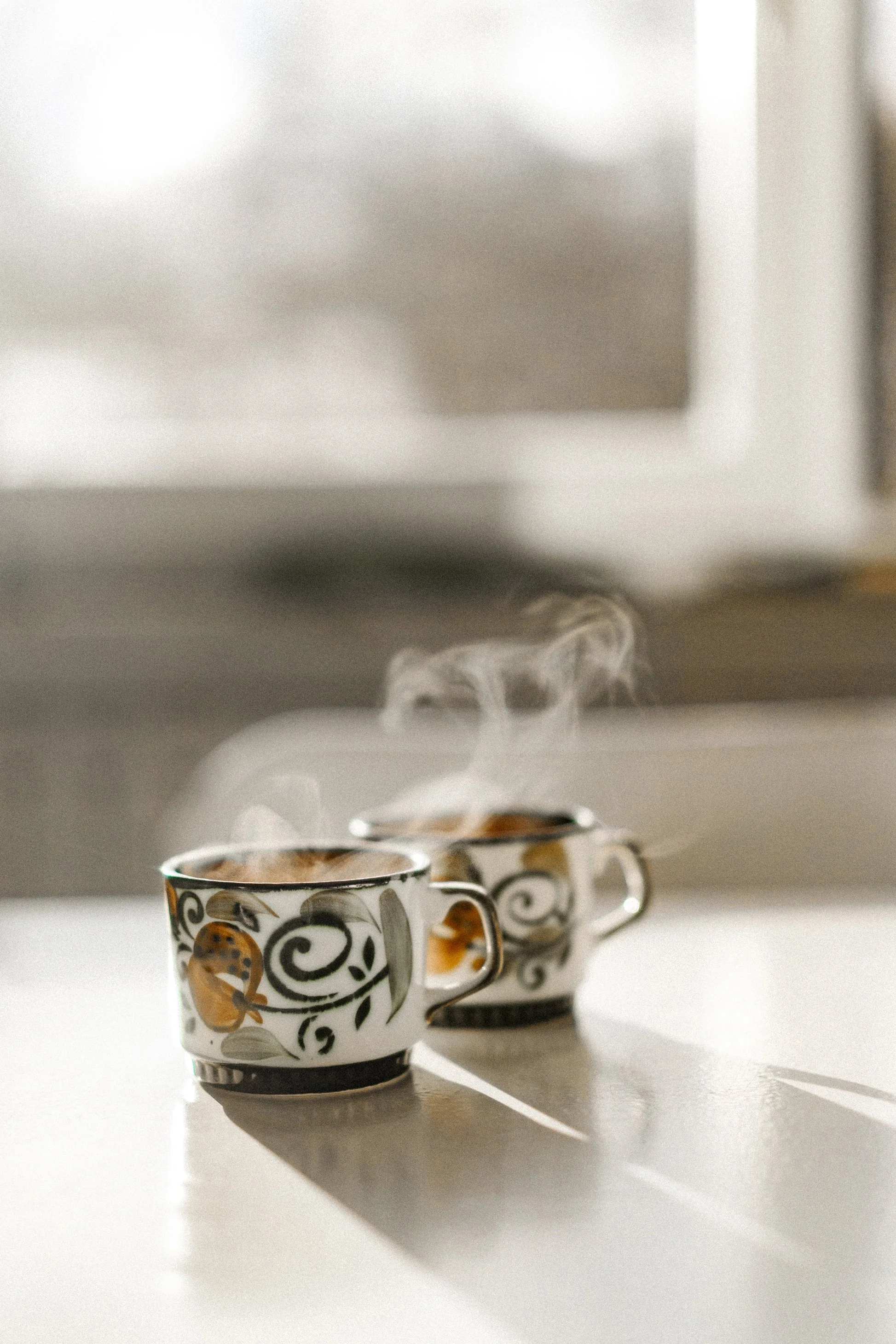 Deux tasses en céramique avec motifs floraux et ornementaux, placées sur une surface blanche, avec de la vapeur de café ou de thé s'élevant au-dessus.