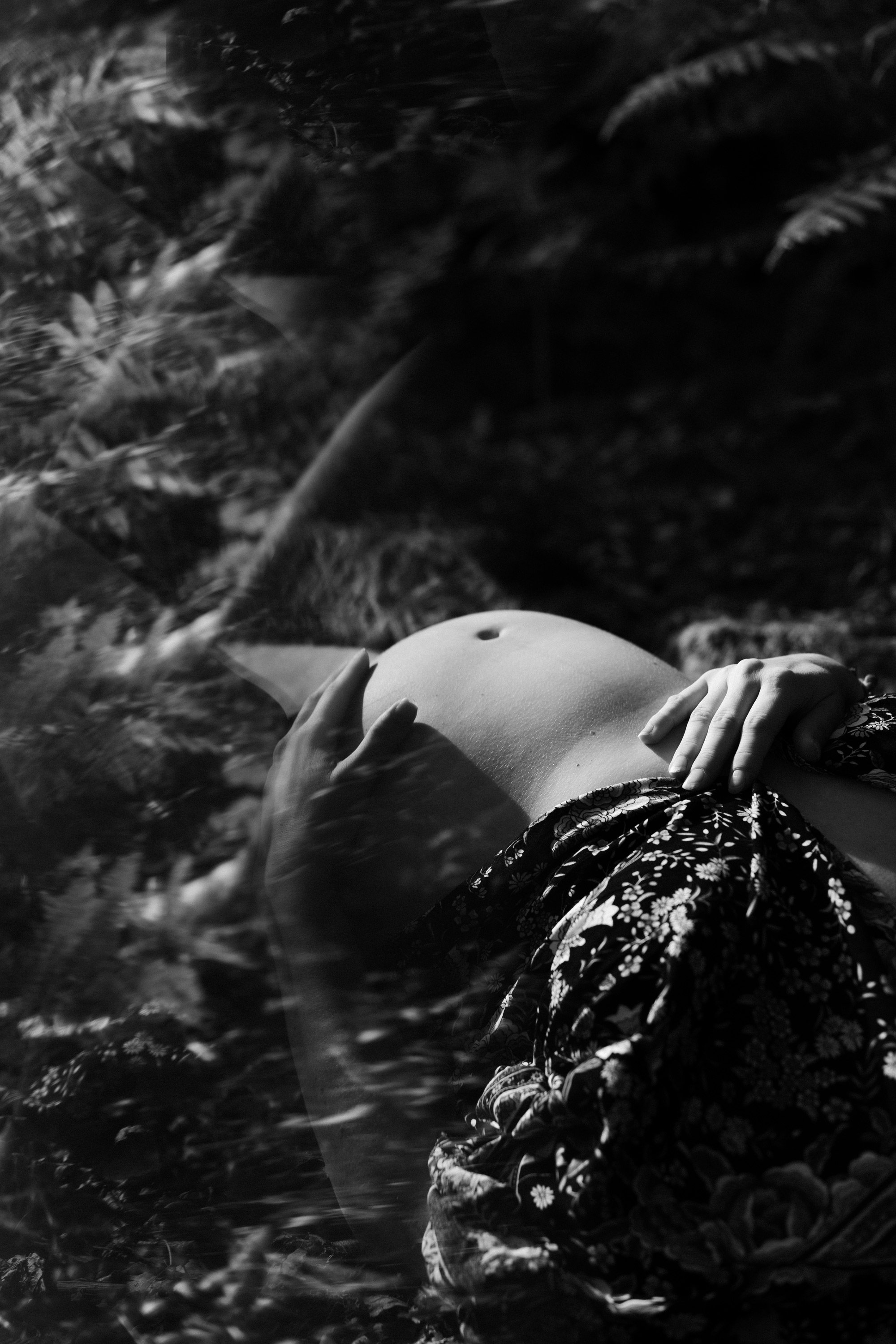 Une personne allongée sur un sol herbeux, portant une jupe à motifs floraux, la main sur l'abdomen, avec de l'eau ou une surface brillante devant elle.