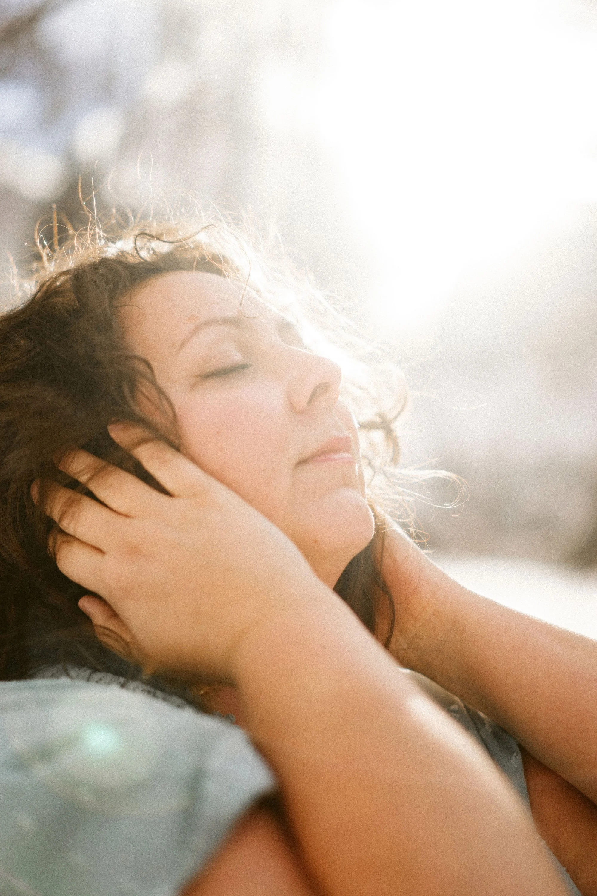 Jeune femme aux yeux fermés, caressant ses cheveux longs et bruns, en lumière naturelle douce.