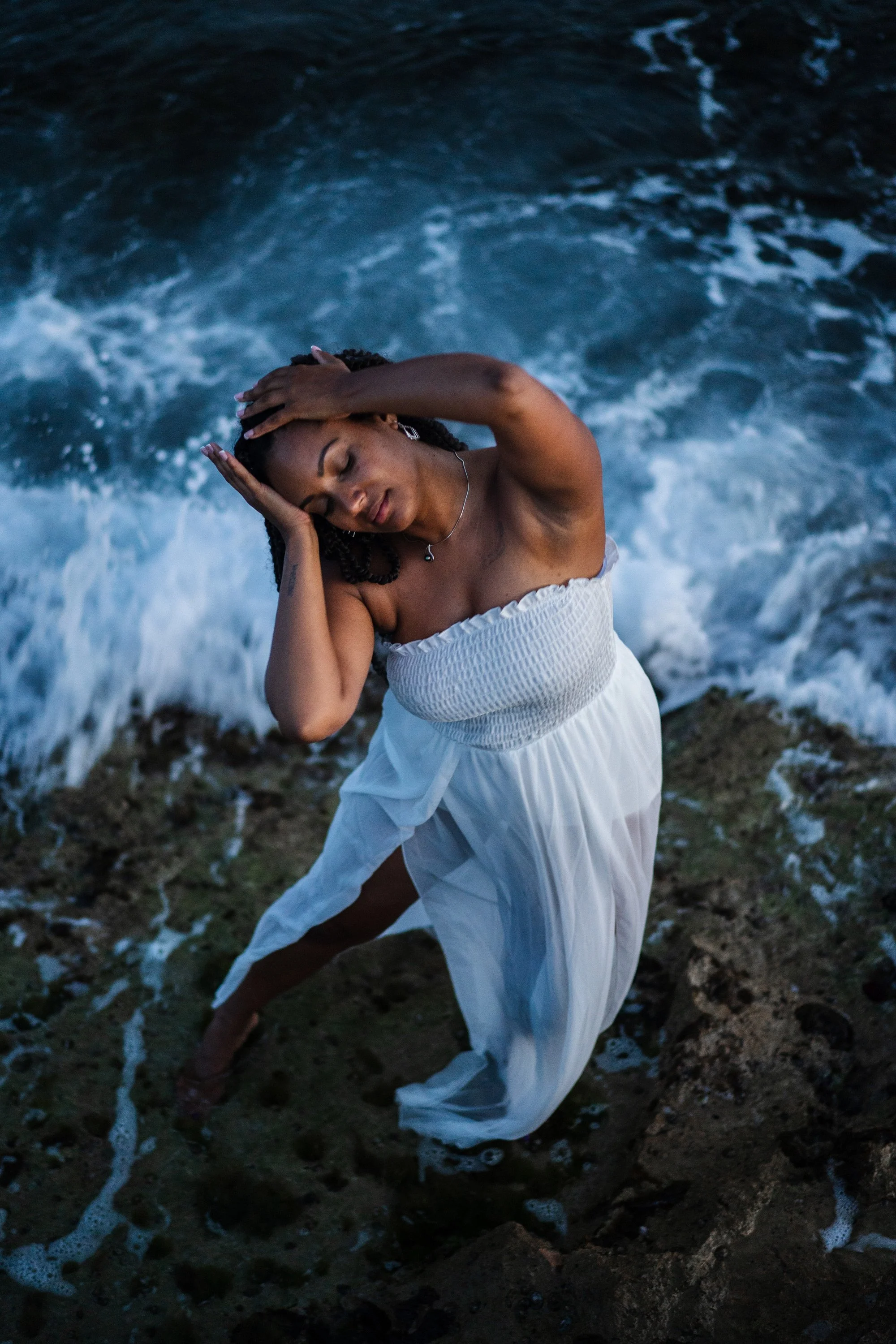 Une femme en robe blanche se tient sur la plage près de la mer, avec ses yeux fermés et une expression détendue.