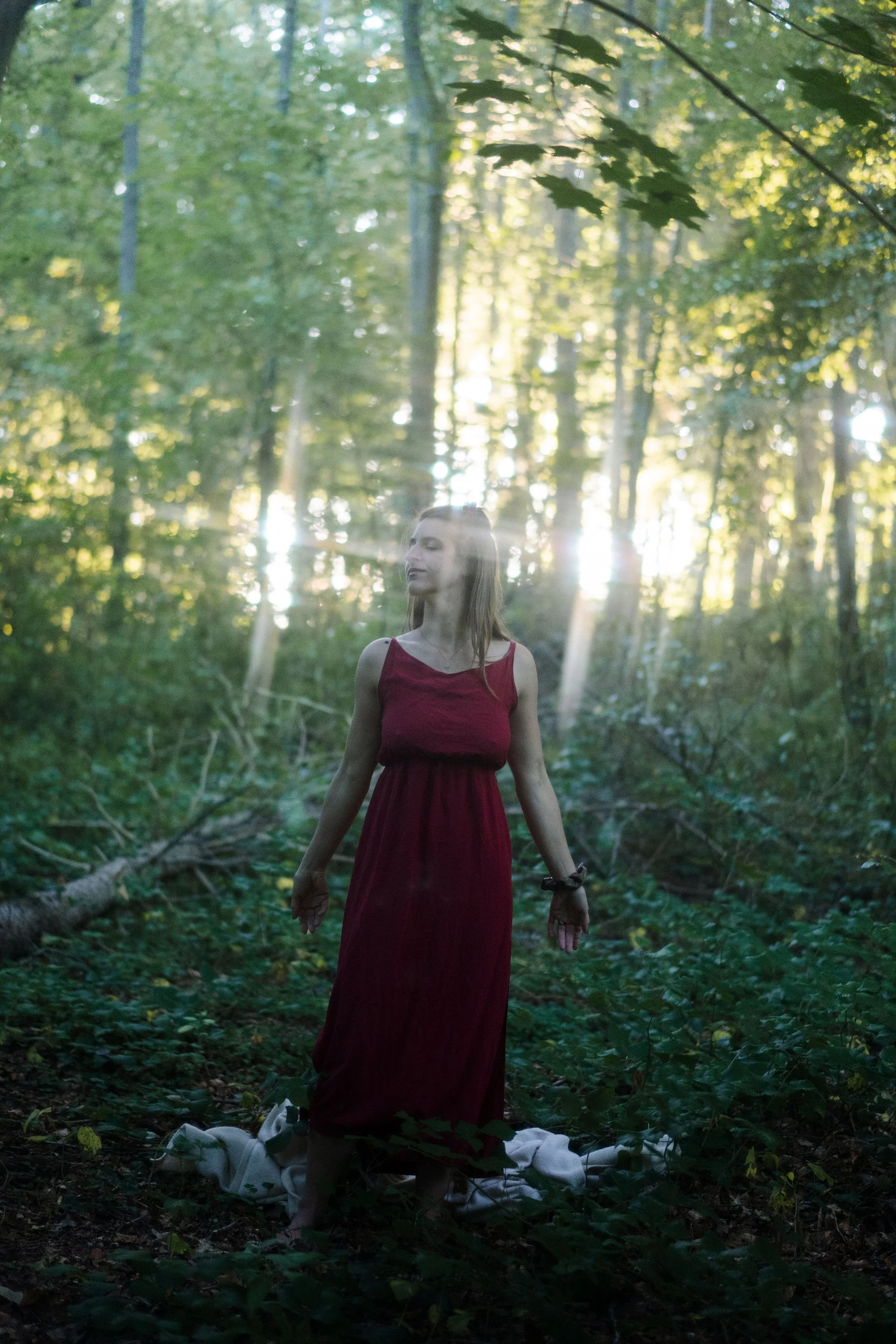 Femme en robe rouge, debout en forêt, avec rayons de soleil à travers les arbres, posé sur un drap blanc au sol.