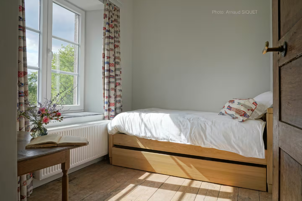 Chambre lumineuse avec un lit en bois, une fenêtre avec vue sur la nature, une table en bois avec un vase de fleurs, et des rideaux à motif.
