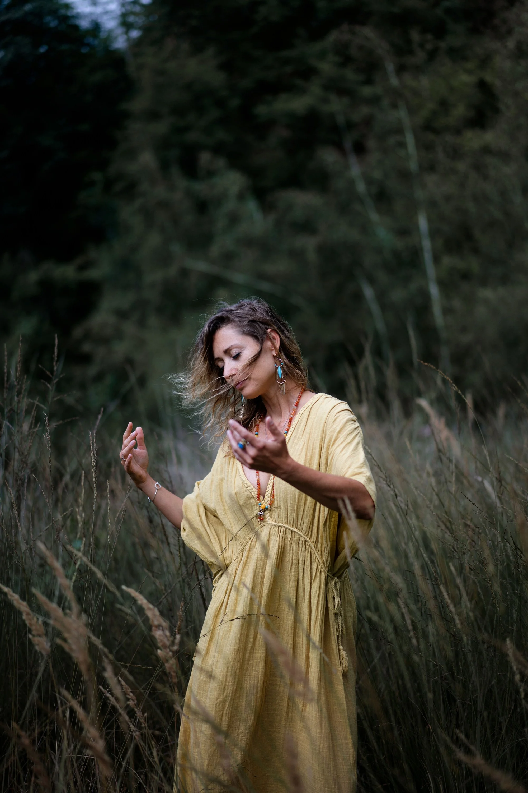 Une femme portant une robe jaune dans un champ d'herbes hautes, avec une végétation dense en arrière-plan, les yeux fermés et les mains levées.