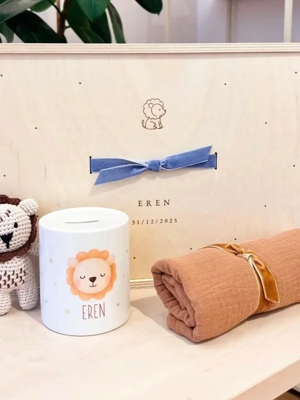 Ein Holzspardose mit Löwenmaske-Motiv, daneben eine Kerze mit Löwenzeichnung und Name Eren, sowie ein gepolstertes Babydeckenrollen, alles auf einem weißen Oberflächen, im Hintergrund ein weißes Möbelstück mit blauer Schleife und ein kleines handgestricktes Tier.