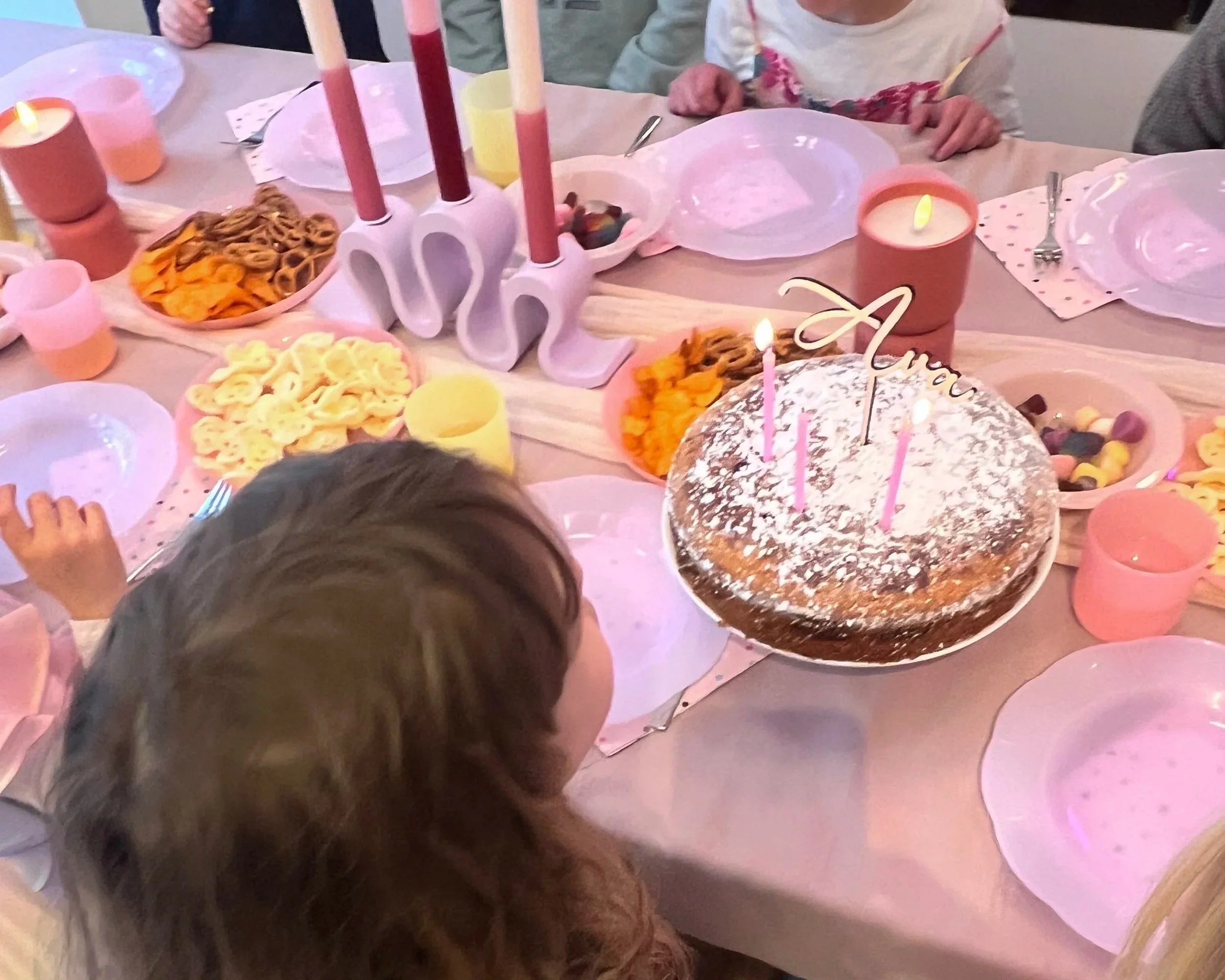 Tisch mit Geburtstagskuchen, Kerzen, bunten Papptellern, Trinkbechern und Snacks, umgeben von mehreren Kindern bei einer Geburtstagsfeier.