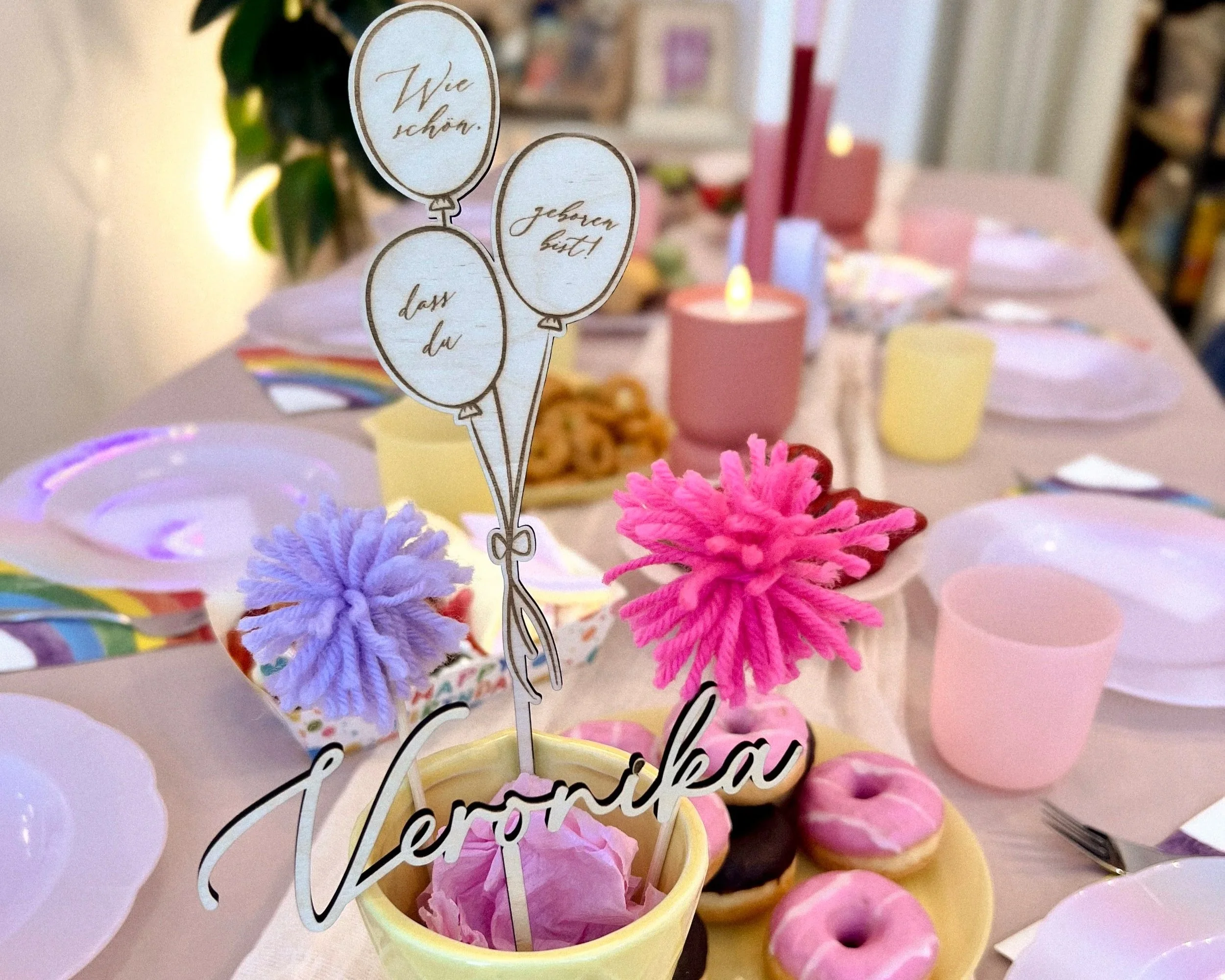 Bunte, geknüpfte Pompons in rosa, lila und rot, dekorativ um einen Kuchen herum, mit einem Kuchenständer in der Mitte. Im Hintergrund sind pinkfarbene Kerzen und Essgeschirr sichtbar.