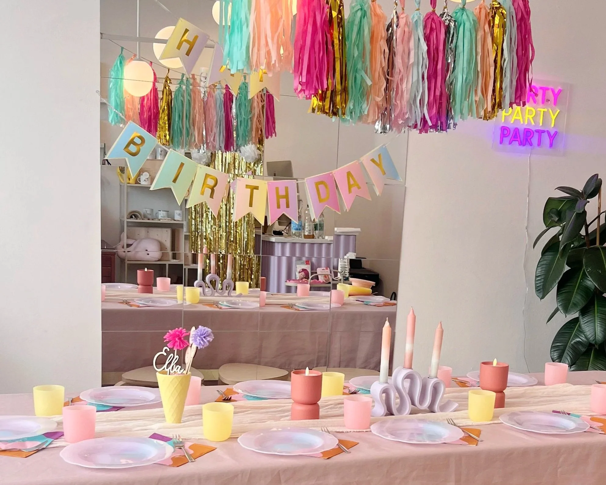 Eine festlich dekorierte Geburtstagstafel mit bunten Kerzen, Pappbechern, Papptellern, Besteck und Dekorationen in Pastellfarben. Im Hintergrund bunte Papier- und Tüll-Streamers sowie eine Lichterkette und eine Neon-Schrift, die 'Party' sagt.