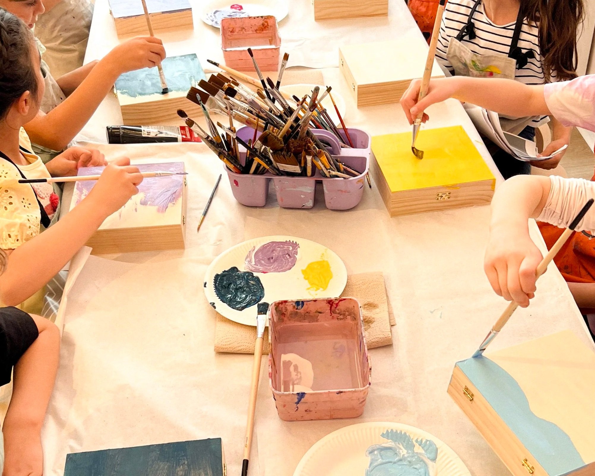 Kinder beim Malen mit Pinseln und Farben, Tische mit Wasserbehältern und Farbtuben, kreative Kunstaktivität in einer Gruppe