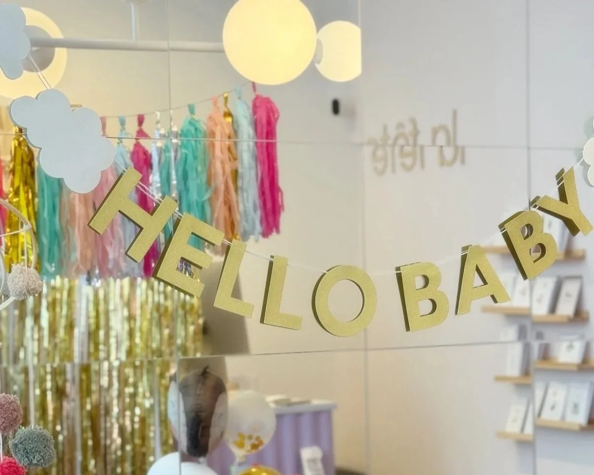 Dekorativer Geburtstagsschmuck mit dem Satz "HELLO BABY" in goldenen Buchstaben, bunte Stoffstreifen und Wolkenformen aus Papier, an einer Wand in einem festlich dekorierten Raum.
