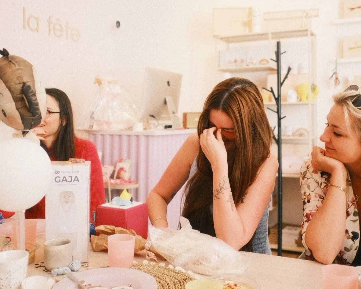 Drei Frauen sitzen an einem Tisch und lachen gemeinsam bei einer Feier, vermutlich bei einer Babyparty, umgeben von Dekorationen und Geschenken.