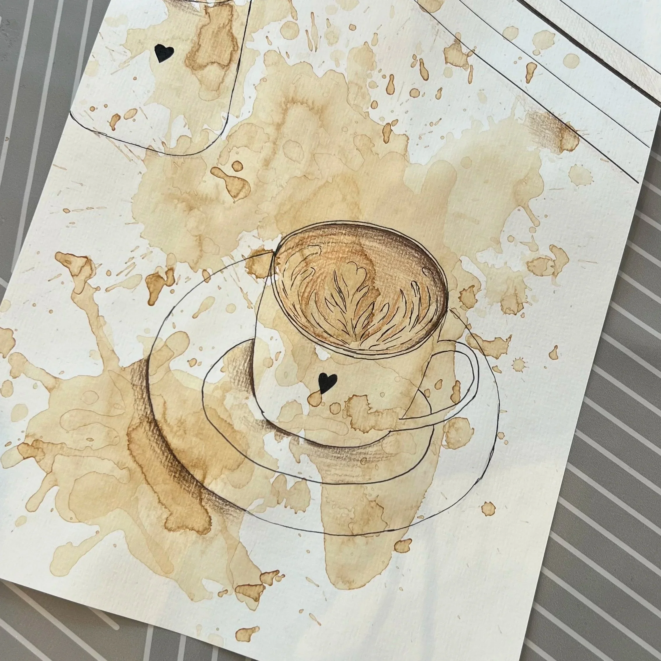 Verwaschene Farbflecken auf einem weißen Blatt Papier, teilweise umrandet mit schwarzen Linien und kleinen schwarzen Herzen, auf einem grauen, gestreiften Untergrund.