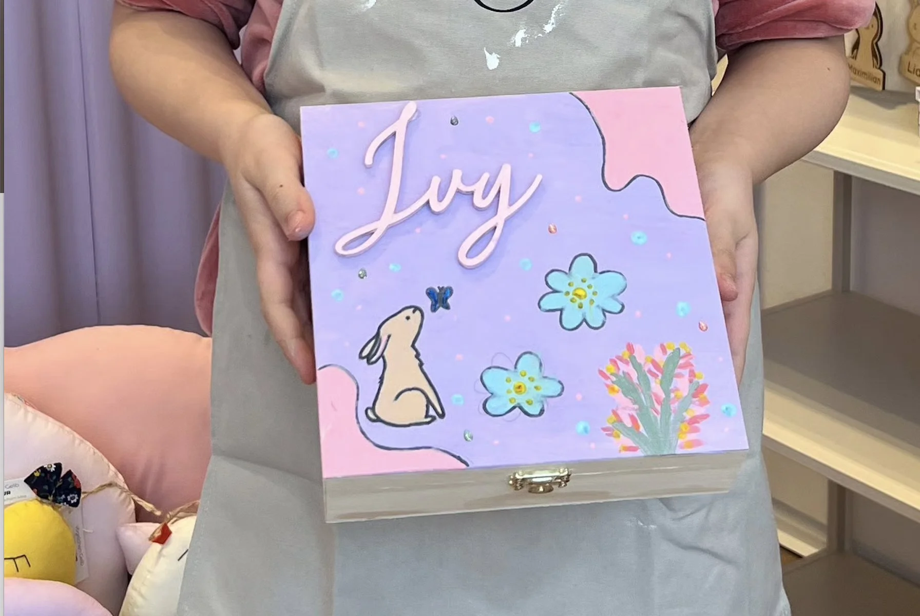 Ein Mädchen hält eine bunte, handgemachte Glückwunschkarte mit dem Namen 'Lysi' und fröhlichen Zeichnungen von Blumen, einem Hasen und einem Schmetterling. Die Karte ist in Pastellfarben gehalten, hauptsächlich Lila und Rosa.