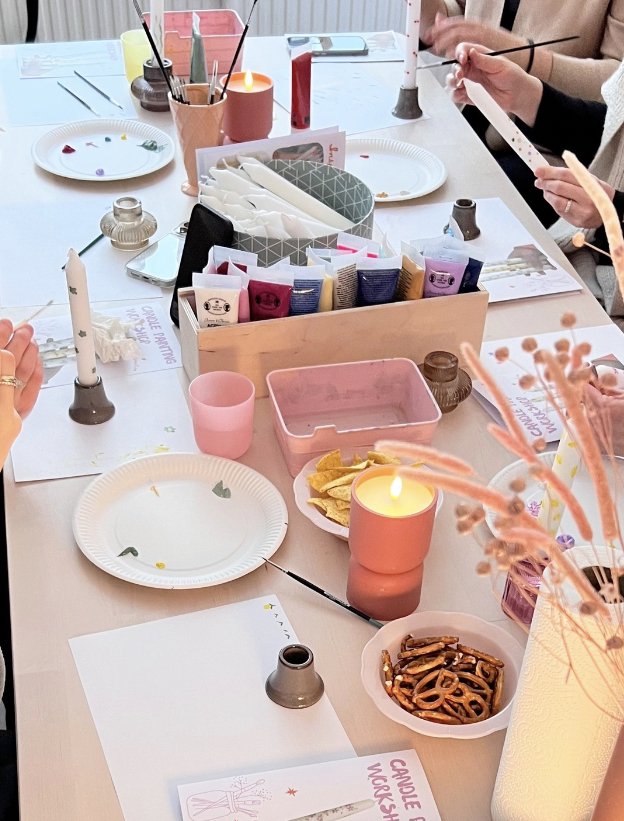 Tisch mit Papptellern, Kerzen, Bastelmaterialien, Snacks wie Brezeln und Chips, und Personen, die basteln oder bastelzubehör verwenden.