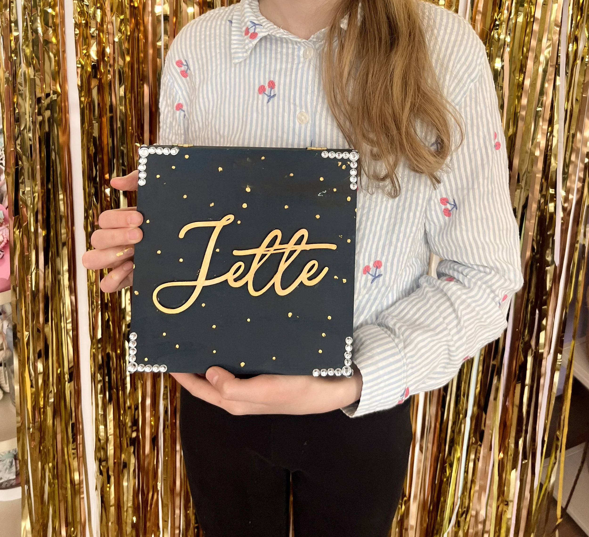 Frau hält eine schwarze Postkarte mit goldener Schrift und weißen Dekosteinen vor sich. Hinter ihr hängen goldene Metallic-Dekorationsstreifen.