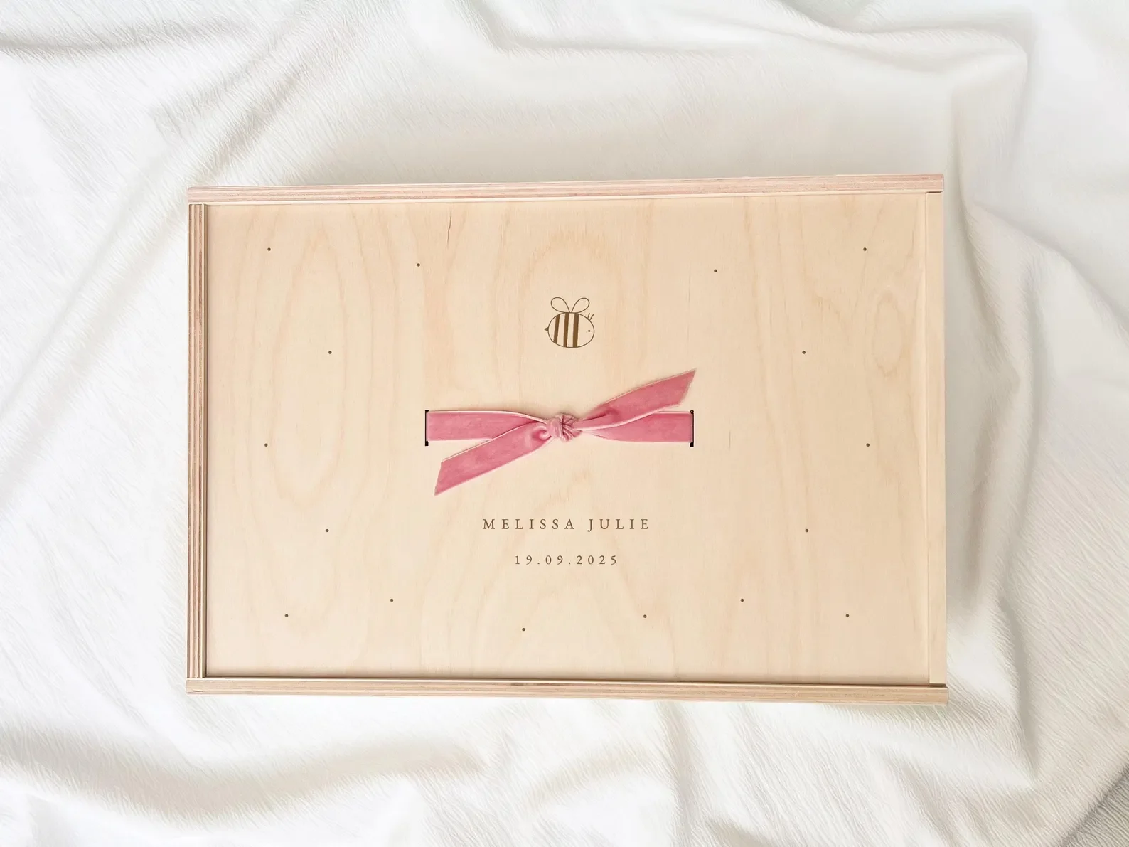 Ein Holzkasten mit einem rosa Schleifenband, das sich in der Mitte knotet, und einer kleinen Biene als Dekoration. Der Text auf dem Kasten lautet: MELISSA JULIE, 19.09.2025.