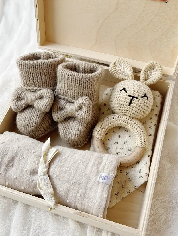 Box mit handgefertigten Babyartikeln wie gestrickte Schuhe, eine gehäkelte Bunny-Ohren-Mütze, ein Ringhalter, ein Stofftuch.