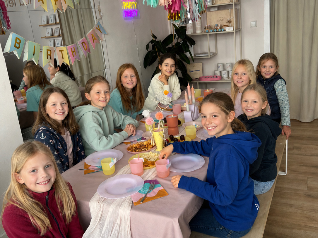 Gruppe von jungen Mädchen sitzen um einen Geburtstagstisch mit Dekoration, Kerzen und Snacks, in einem Festsaal mit bunten Luftschlangen und einem 'Happy Birthday'-Banner.