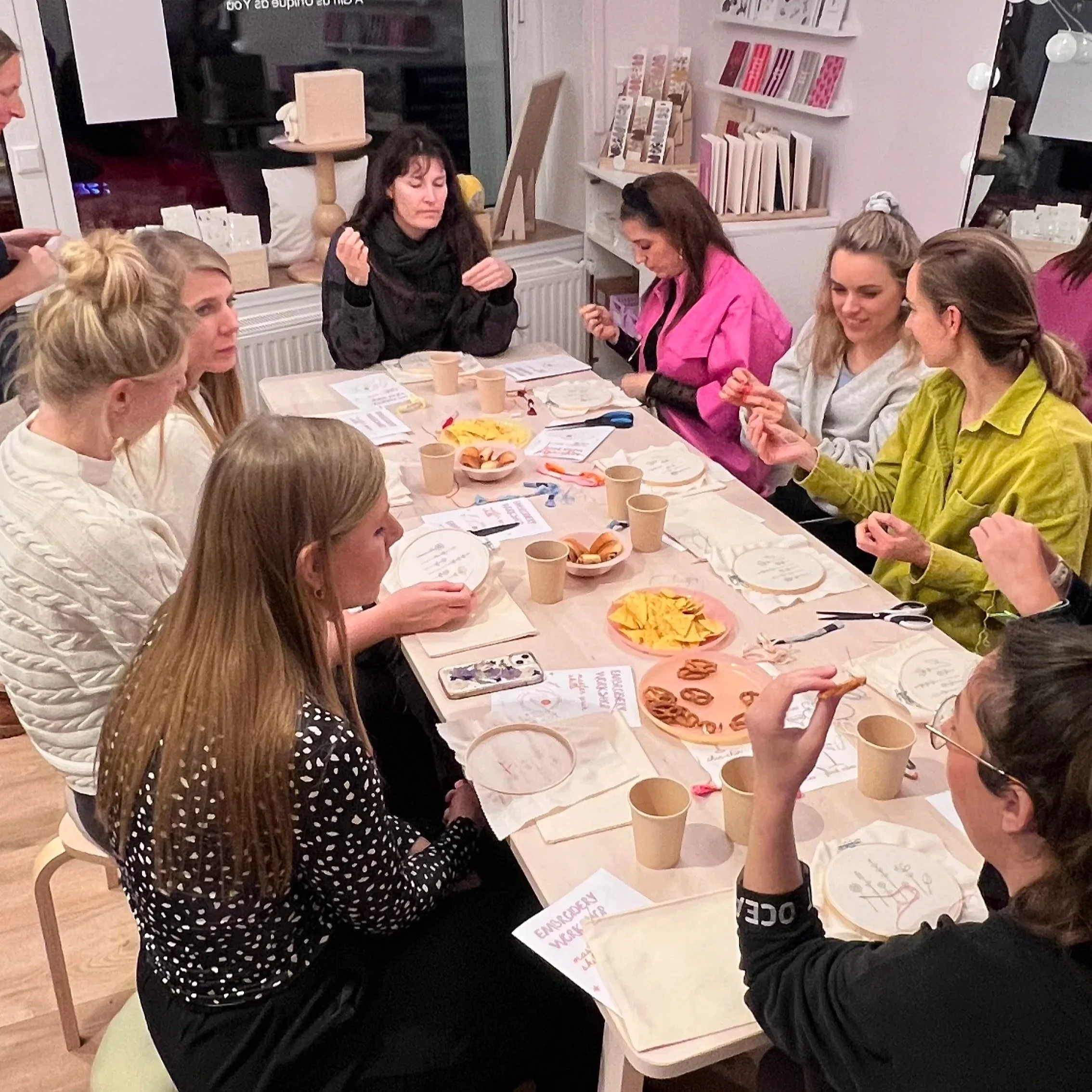 Mehrere Frauen sitzen um einen Tisch in einem Laden, der mit Büchern und Schreibwaren dekoriert ist, beim gemeinsamen Essen.
