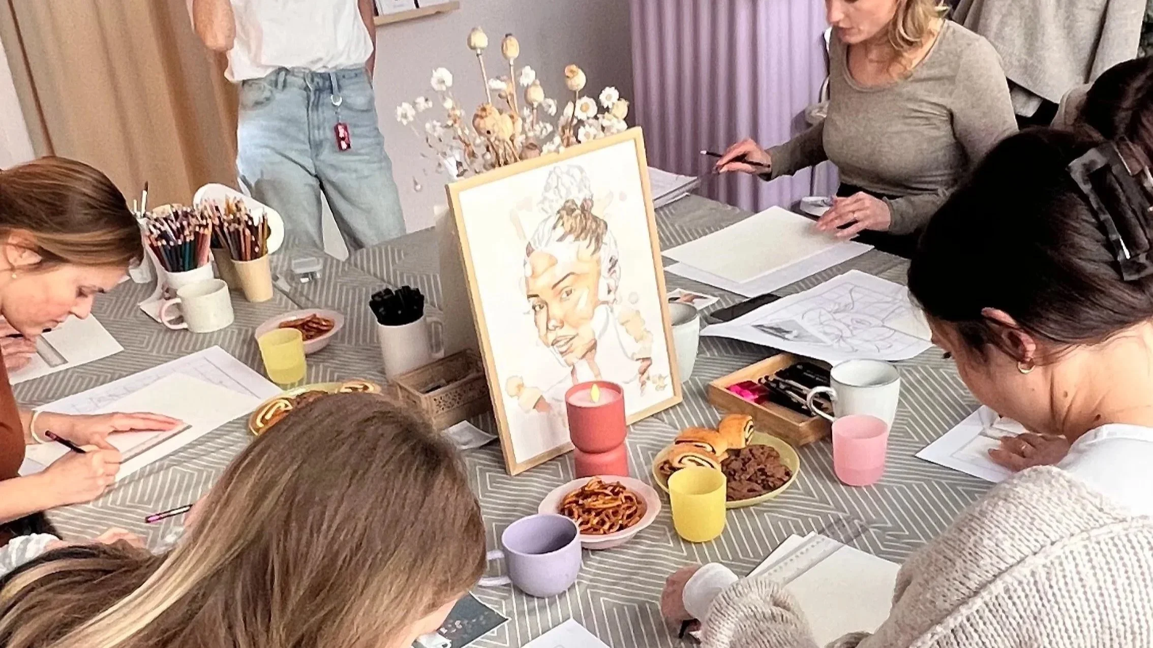 Gruppe von Menschen, die an einem Tisch beim Malen und Zeichen beteiligt sind, mit Zeichenmaterialien, Desserts und einem gemalten Porträt aufgestellt.