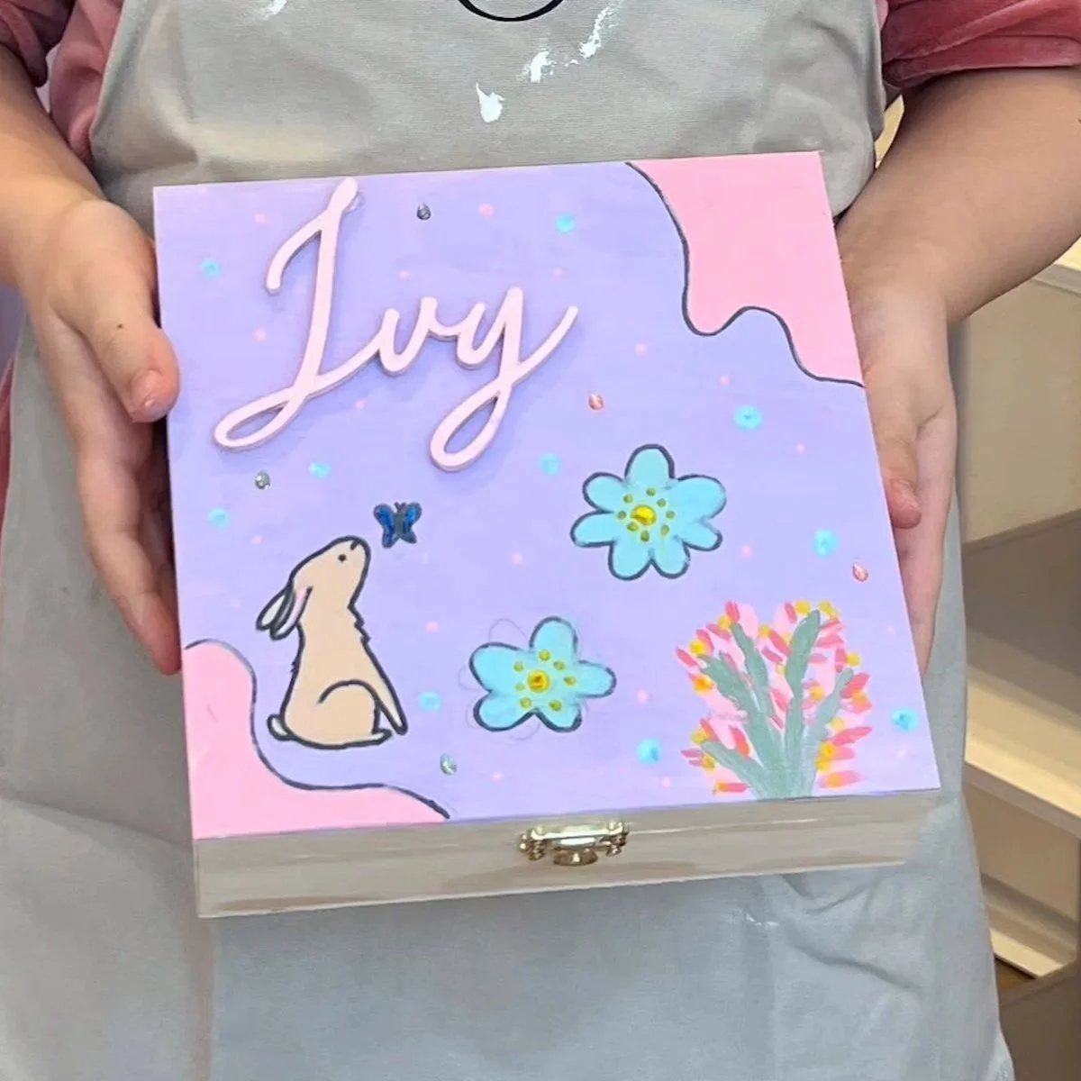 Eine Hand hält eine bunte Schachtel mit dem Namen 'Lizzy' auf der Oberfläche, verziert mit einem Häschen, Blümchen, einem Schmetterling und einer Blume, alles in Pastellfarben gestaltet.