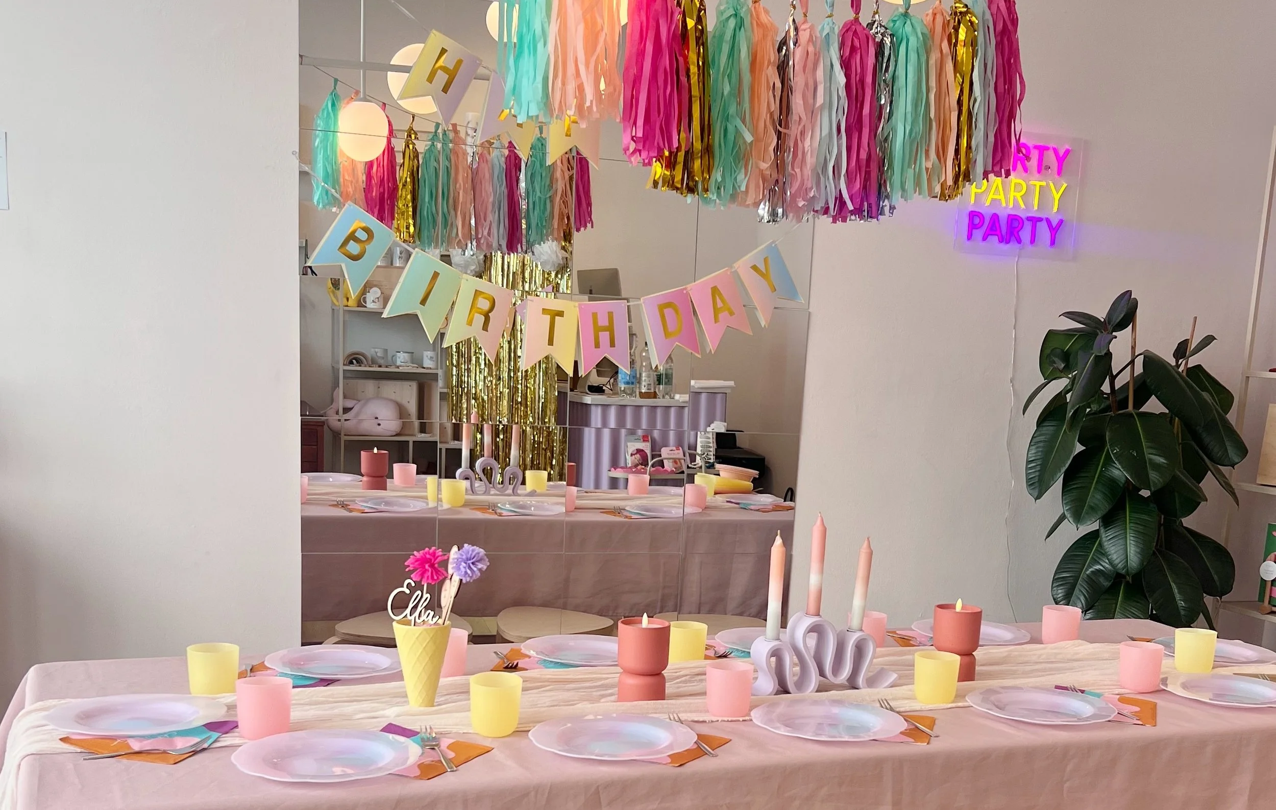 Ein bunt dekorierter Raum für eine Geburtstagsfeier mit Papiergirlanden, Kerzen, Geschirr und einem lange gedeckten Tisch. Im Hintergrund sind bunte Pompons, eine Tafel mit Laternen und eine Neon-Schrift, die 'Party' sagt.
