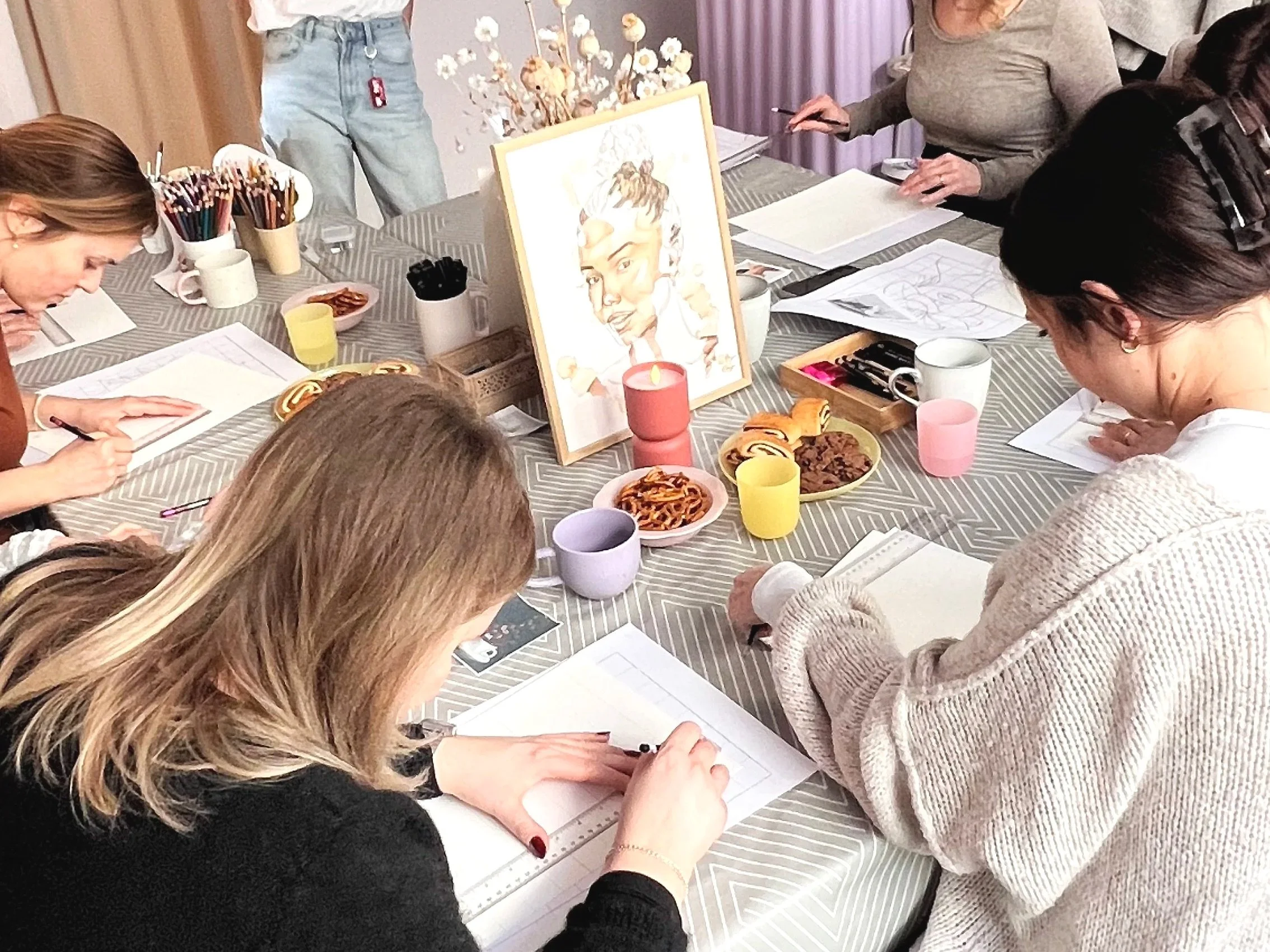 Mehrere Frauen sitzen um einen Tisch, zeichnen und schreiben auf Papier. Der Tisch ist mit Süßigkeiten und Trinkgefäßen gedeckt, es gibt eine Frischlingszeichnung auf einem Stehbild im Hintergrund.
