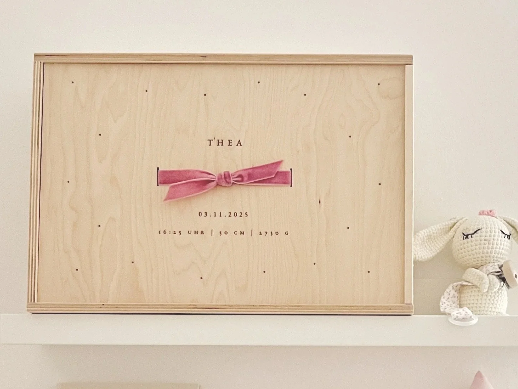 Holzkistchen mit Geburtsdaten des Babys: Name THEA, Geburtsdatum 03.11.2025, Uhrzeit 16:25 Uhr, Größe 50 cm, Gewicht 2770 g, mit pinkem Band. Neben der Kiste ein gehäkeltes Kuscheltier mit geschlossenen Augen.
