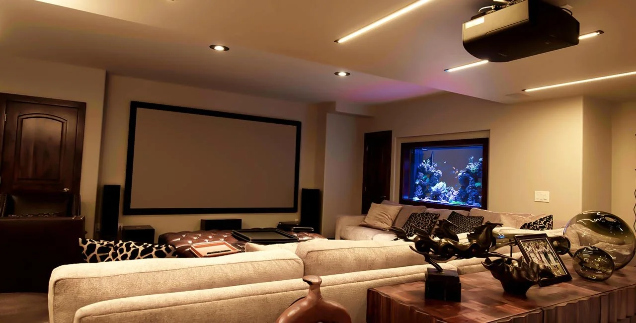 Living room with a large beige sectional sofa, a fish tank on the wall, a ceiling projector, and decorative items on a wooden table.