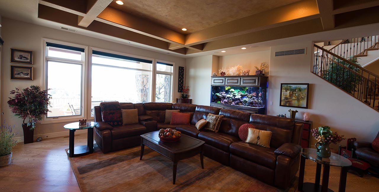 Living room with large windows, brown leather sectional sofa adorned with various pillows, a wooden coffee table with a fruit bowl, an aquarium on the wall, potted plants, and a staircase on the right side.