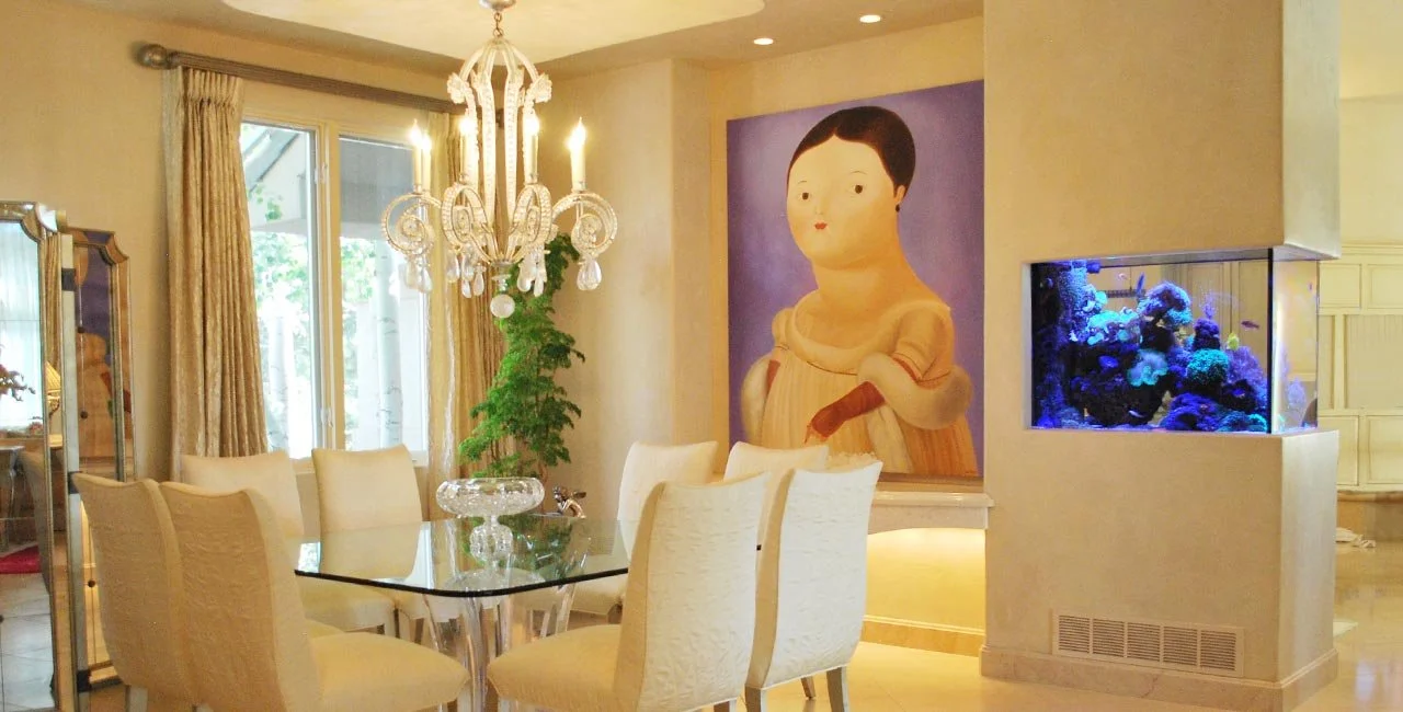 An elegant dining room with a glass table surrounded by cream-colored chairs, a chandelier hanging above, large windows with curtains, a potted plant, a large portrait of a young child on the wall, and a built-in aquarium with blue lighting.