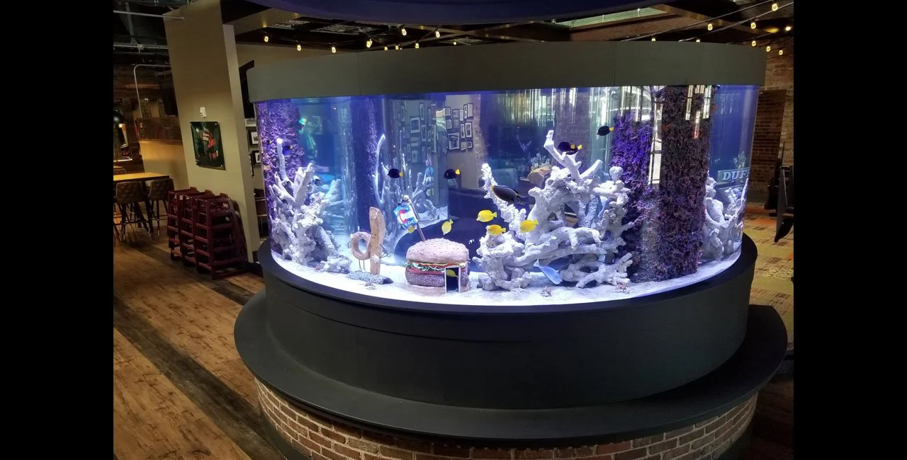 Large oval saltwater aquarium with colorful fish, coral, and reef decorations inside a cozy indoor setting, possibly a restaurant or cafe.