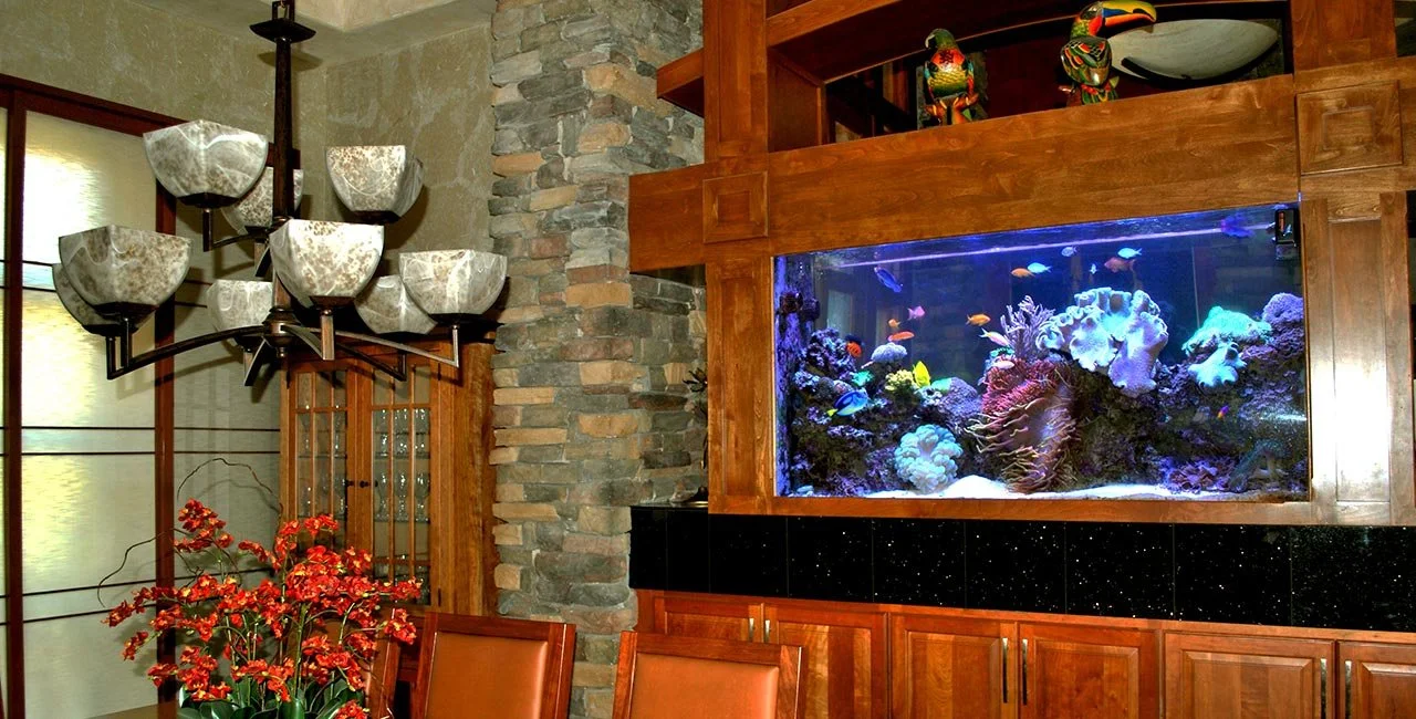 Living room with stone fireplace, large built-in saltwater aquarium with fish, wooden cabinetry, and decorative parrot statues on top of the aquarium.