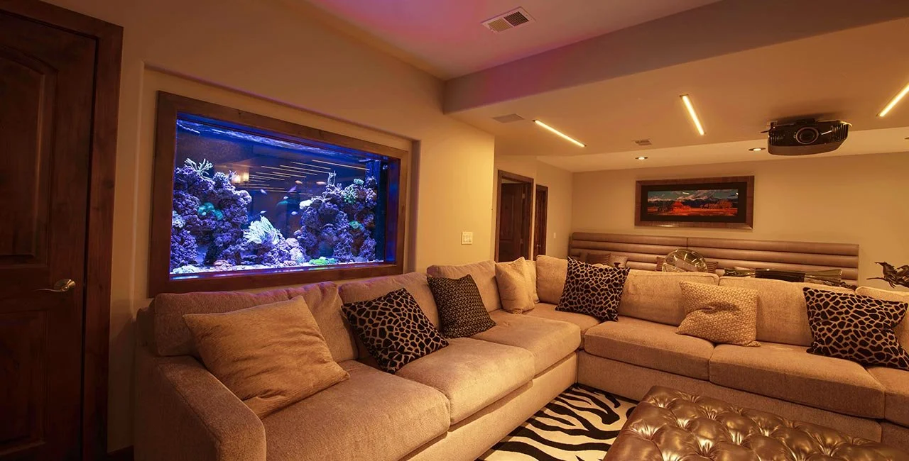 Living room with a large sectional sofa decorated with various patterned and solid-colored pillows, an illuminated saltwater aquarium built into the wall, a zebra-striped rug, a framed picture on the back wall, and ceiling lights, with a projector mo