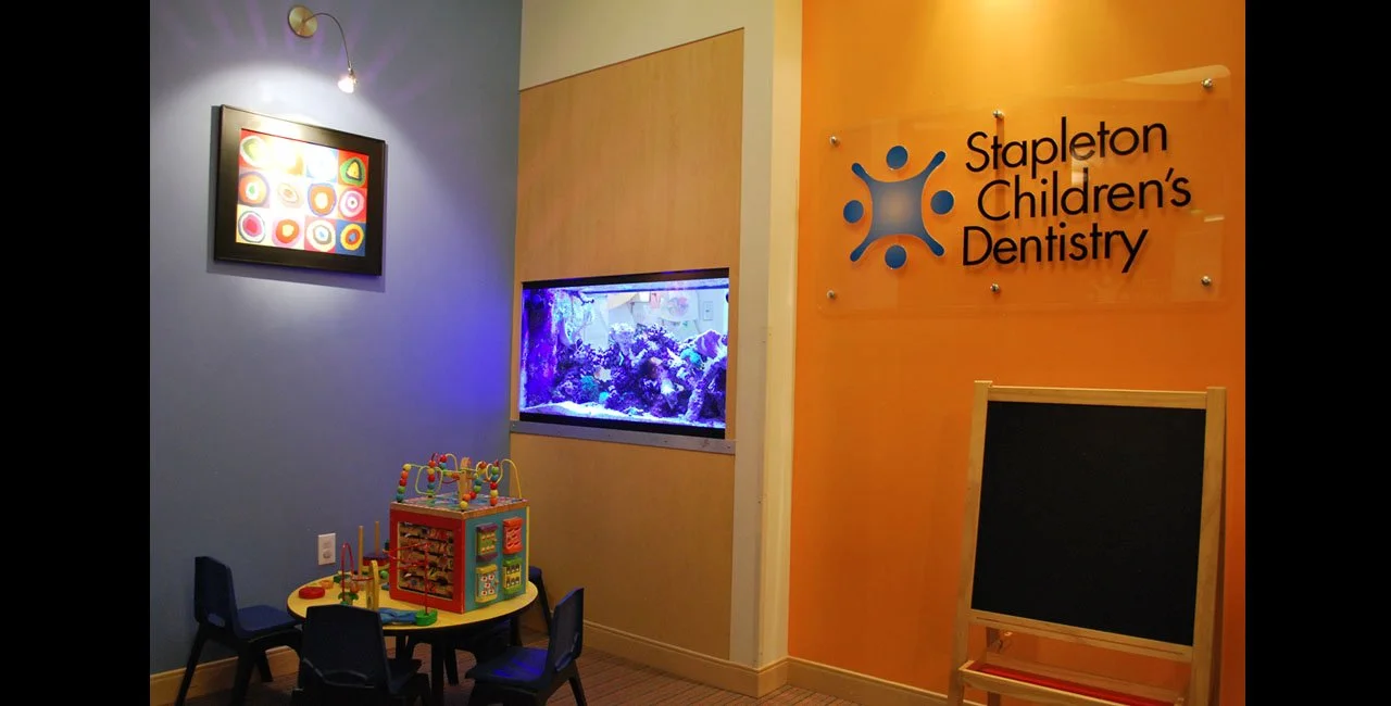 Interior of a children's dental office with a blue wall, a framed colorful artwork, a fish tank, and a Kids' activity table with toys. There's also an orange wall with a sign for Stapleton Children's Dentistry and a black chalkboard easel.