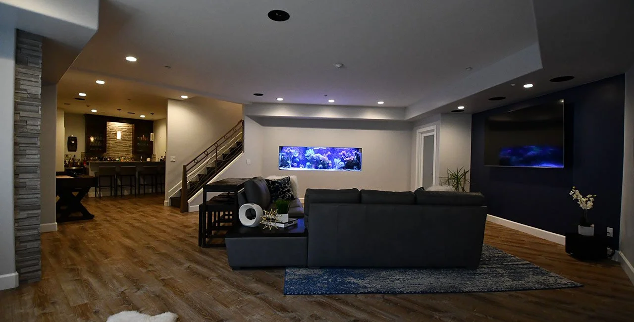 Modern living room with wooden floors, a gray sectional sofa, a large fish tank on the wall, a flat-screen TV on a dark accent wall, and a small plant in a black pot.