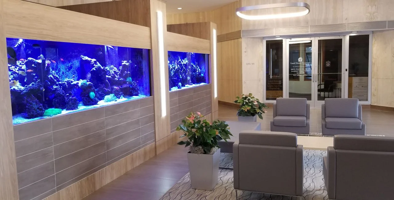A modern lobby with two large saltwater aquariums, potted plants, four gray armchairs, and sliding glass doors.