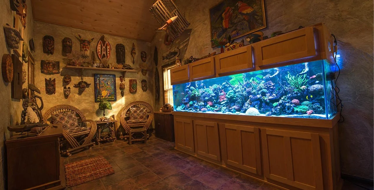 A room with a large saltwater aquarium filled with colorful coral and fish, two rattan chairs with patterned cushions, a small table between them with a lamp, tribal masks and artwork on the walls, a window, and a wooden ceiling.