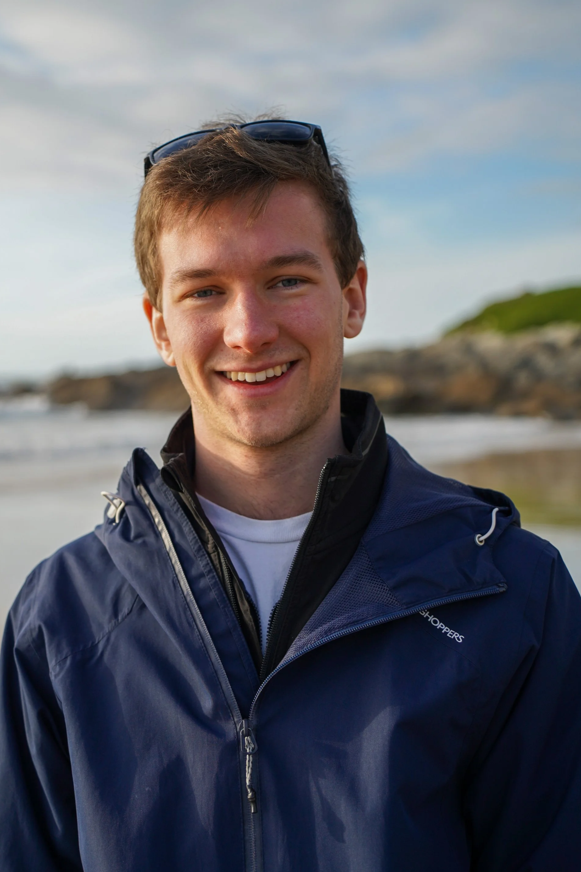 A smiling young man wearing a navy blue riding jacket with sunglasses resting on his head, on a beach with rocky cliffs and cloudy sky in the background.