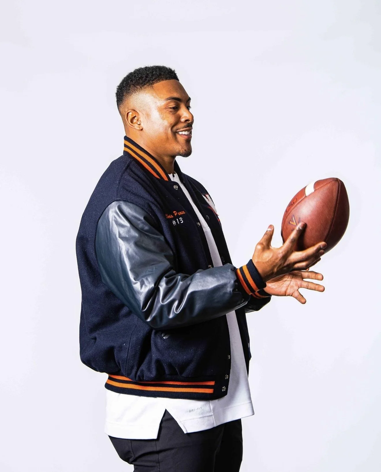 A young man with a short haircut holding a football while wearing a navy blue varsity jacket with leather sleeves, smiling, against a light background.