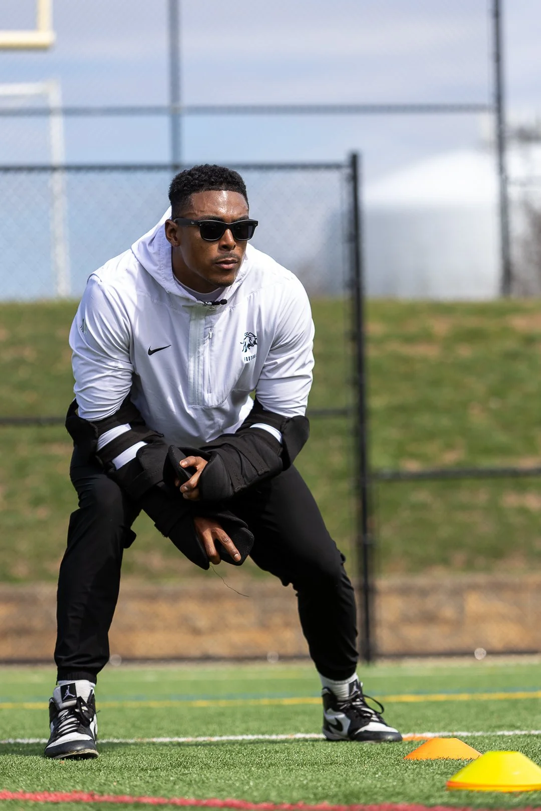 A male athlete in sunglasses and sportswear crouching on a sports field during training, with cones set up in front of him.