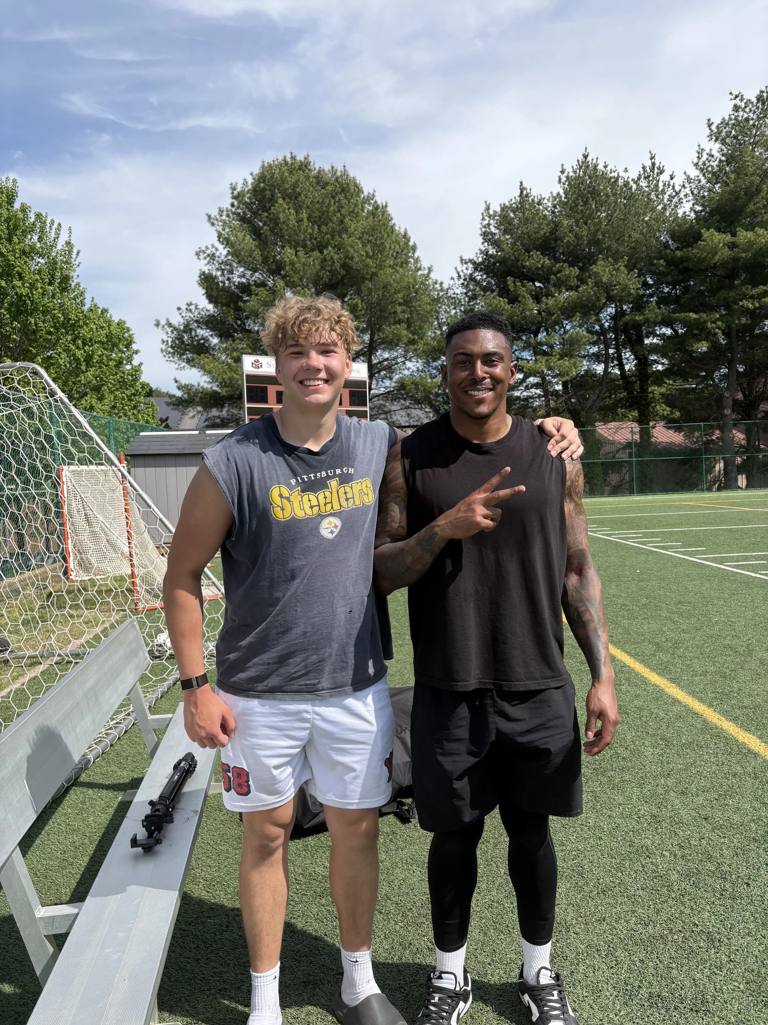 Two young men standing outdoors on a sports field, smiling at the camera, with trees and a scoreboard in the background.