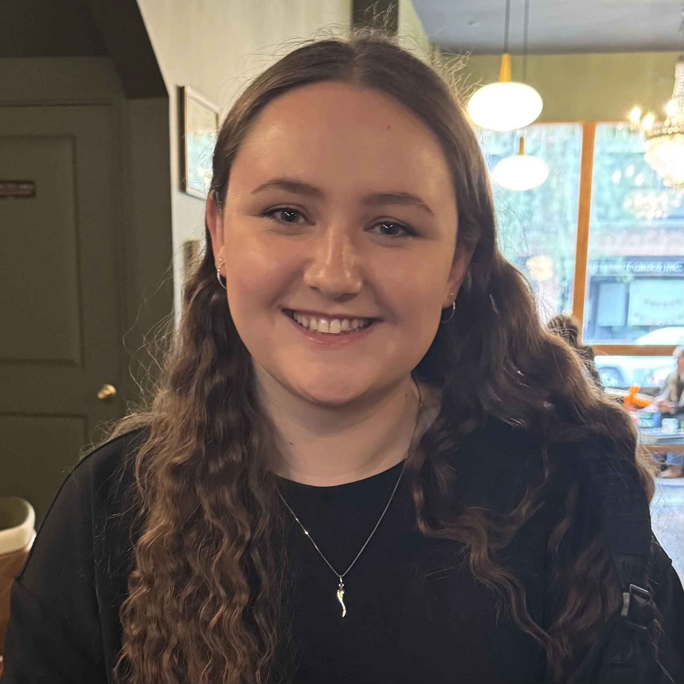 Headshot of smiling female with long, wavy brown hair in a black shirt with a silver necklace..