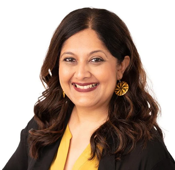 Headshot of smiling female with medium-length, curly brown hair in a black blazer, yellow shirt, and gold earrings..